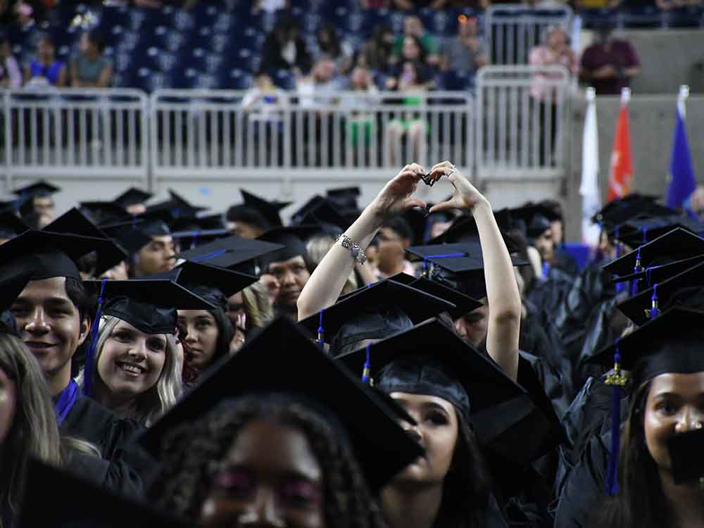 San Jacinto College graduate in cap and gown making a heart gesture with her hands raised in the air during the graduation ceremony